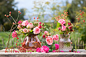 Various bouquets of roses (pink) and rose hips in glass vases on wooden table in garden