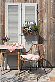 Rattanstuhl und Tisch vor Holzwand mit dekorativen Fensterläden und Blumenarrangement