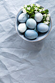 Blaue Ostereier und weiße Blüten in Keramikschale auf blauer Leinentischdecke