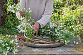 Gesteck aus einem gebundenem Kranz aus Klematisranken (Clematis) in Schale mit Wasser, mit Myrtenaster (Aster ericoides) und Hagebutten