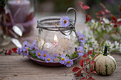 Kerze im Glas dekoriert mit Blüten der Myrtenaster (Aster ericoides), und Silberblatt, Herbstlaub und Zierkürbis auf Gartentisch