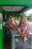 Ein Schweinekopf und Fleisch an Fleischhaken in einer Freiluftmetzgerei in Bonao, Dominikanische Republik. Der Metzger schneidet den Kadaver im Hintergrund auf, während zwei Kunden warten.