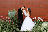 Groom kissing brides hand against brick wall and flowers