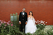 Portrait of smiling bride and groom against brick wall and flowers
