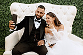 Smiling bride and groom sitting on sofa, taking selfie