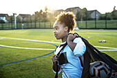 UK, Rückansicht einer Fußballspielerin (12-13), die eine Tasche mit Bällen auf einem Feld trägt