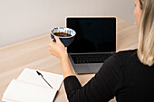 Close-up of woman holding coffee cup at desk with laptop and notepad