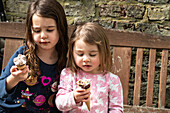 UK, Two girls (2-3, 4-5) with ice cream cones on bench