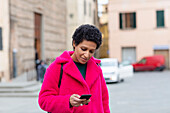 Italien, Toskana, Pistoia, Frau im rosa Mantel benutzt Smartphone auf der Straße