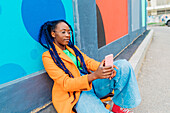 Italien, Mailand, Frau mit Zöpfen sitzt an bunter Wand und benutzt Smartphone