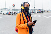 Italien, Mailand, Frau mit Gesichtsmaske und Kopfhörern hält Smartphone auf der Straße