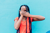Italy, Milan, Young woman covering mouth and eye in front of blue wall