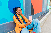 Italy, Milan, Woman with braids sitting by colorful wall, using smart phone