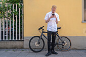 Deutschland, Köln, Albinomann mit Smartphone vor Gebäude und Fahrrad
