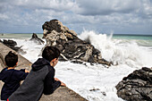 Boys (6-7, 12-13) looking at sea waves crashing against rocks, Sicily, Italy
