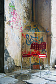 Ein roter Plastikstuhl sitzt neben einer verwitterten Wand mit aufgesprühtem Graffiti; Barcelona, Spanien