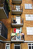 Fahrräder und Wäsche hängen auf kleinen Balkonen an einem Wohnhaus aus Backstein, Portobello Road; London, England.