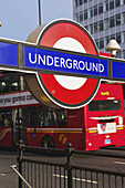 Ein Schild für die Treppe zur U-Bahn neben einer Straße mit einem vorbeifahrenden Doppeldecker-Bus; London, England.
