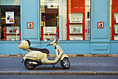 Ein motorisierter Roller, geparkt vor einem blauen Gebäude entlang einer Straße, Canal Saint Martin; Paris, Frankreich.