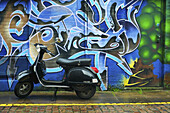 Ein motorisierter Roller, der vor einem Gebäude mit abstrakten Kunstwerken an der Wand geparkt ist; Notting Hill; London, England