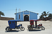Ein roter und blauer dreirädriger Wagen sitzt auf dem Sand vor einem blauen Gebäude mit blauem Himmel und Palmen; Sechura-Wüste, Peru