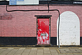 Eine rote Tür mit abblätternder Farbe an einem gestrichenen Backsteingebäude; London, England.