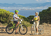 Ein Mann und eine Frau auf Fahrrädern stehen mit Blick auf das Meer und die Küste; Tarifa, Cádiz, Andalusien, Spanien
