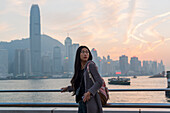 Eine junge Frau am Hafen mit Blick auf die Skyline bei Sonnenuntergang, Kowloon; Hongkong, China.