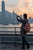 A Young Woman Takes A Picture With Her Camera Of The Harbour And Hong Kong Skyline At Sunset, Kowloon; Hong Kong, China