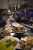 Ein Barmann steht unter hängenden Schinkenbeinen (Jamon Iberico) mit einer Auswahl an Pintxos auf dem Bartresen; San Sebastian, Spanien