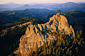 Aerial view of Pilot Rock at twilight. The iconic rock face is a plug of volcanic basalt that juts 400 feet above Cascade–Siskiyou National Monument in a crossroads of mountain ranges, geological eras and habitats. The 65,000-acre monument is at the junction of the Oregon and Cascades and Siskiyou Mountains with Mt. Shasta on the left rising in the far distance across the state line in California; Oregon, United States of America