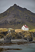 Einsames weißes Haus und zerklüfteter Berg mit Lavafelsen entlang der Meeresküste bei Arnastapi, Snaefellsnes, Westisland; Arnastapi, Island.