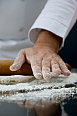 Close up of a chef hand covered in flour rolling a rolling pin across a dough over a glass table