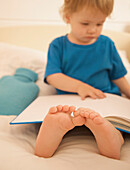 Junge sitzt im Bett mit einem aufgeschlagenen Buch über seinen Beinen und Füßen