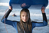 Lächelndes und nasses Mädchen, das ein Surfbrett auf dem Kopf hält und aus dem Meer kommt