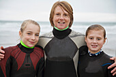 Frau und zwei Mädchen in Surfanzügen stehen lächelnd am Strand