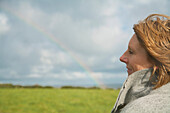 Nahaufnahme einer Frau, die auf einem Feld steht und einen Regenbogen betrachtet