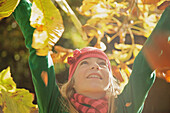 Nahaufnahme eines lächelnden jungen Mädchens, das seine Arme in die Luft streckt und von Blättern umgeben ist
