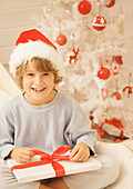 Boy sitting by Christmas tree in a red and white furry hat unwrapping a gift