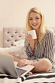 Lächelnde junge Frau, die auf dem Bett sitzt und einen Laptop benutzt