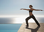 Young woman practicing yoga by a swimming pool with ocean in the background