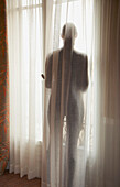 Silhouette einer Frau hinter einem halbtransparenten Vorhang, Rückenansicht
