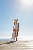 Frau in weißem Hut und Badeanzug steht auf einer Sonnenterrasse und hält ein Handtuch