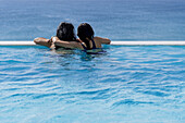 Rückansicht zweier Menschen, die sich umarmen und vom Schwimmbad aus auf das Meer blicken