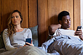 Adult couple lying on bed with upset look on faces while using smart phone