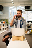 Männlicher Unternehmer, der auf einem Tisch sitzt und mit einem Smartphone telefoniert