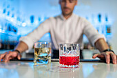 Male bartender serves refreshing whisky and Negroni cocktails in glasses while standing behind the counter and looking at blurred shelves of bottles