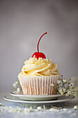 A white chocolate muffin with buttercream, grated white chocolate and a stemmed maraschino cherry. Presented on a plate with white flowers