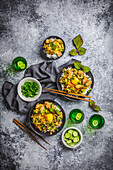 Anrichten von dunklen Schalen mit japanischem Huhn, Ei und Reis, mit grünen Sakebechern und Schale mit Gurkenscheiben