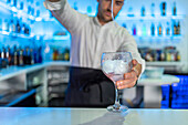Male bartender holding a glass and looking down while pouring a blue coloured cocktail on ice into a transparent glass and spraying it on the bar counter in front of a blurred shelf of bottles